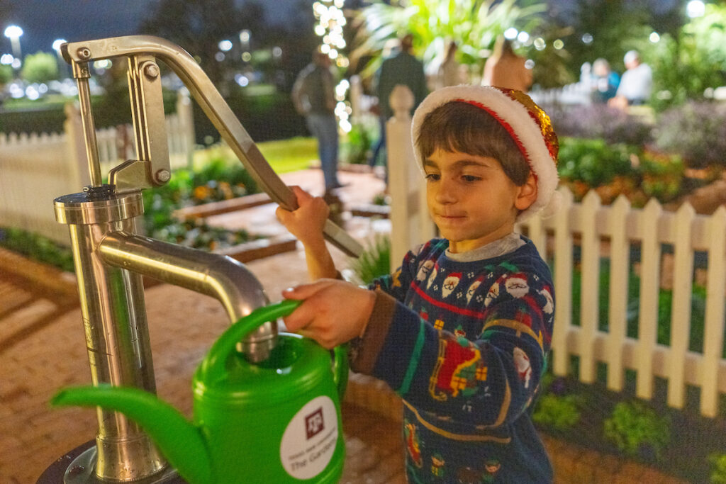 A young child wearing a Santa hat and festive sweater fills a waterig can from a pump at The Gardens at Texas A&M.
