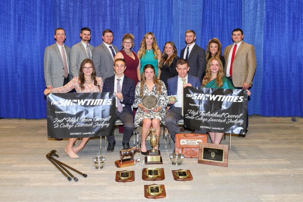 A group of people stand and sit with in nice clothes and hold award banners, with a number of other plaques and trophies at their feet from the national collegiate Livestock Judging competition.