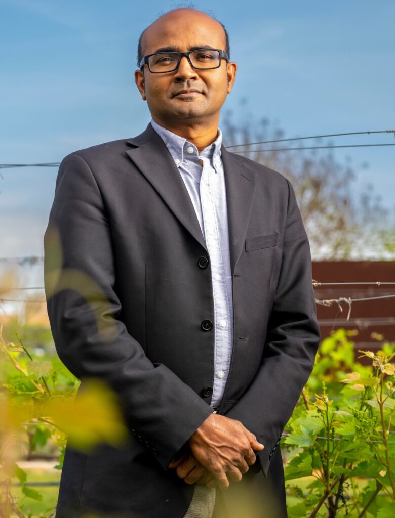 A man in a suit jacket posing in an outdoor field with buildings behind him - Muthu Bagavathiannan earned the Humboldt Research Fellowship