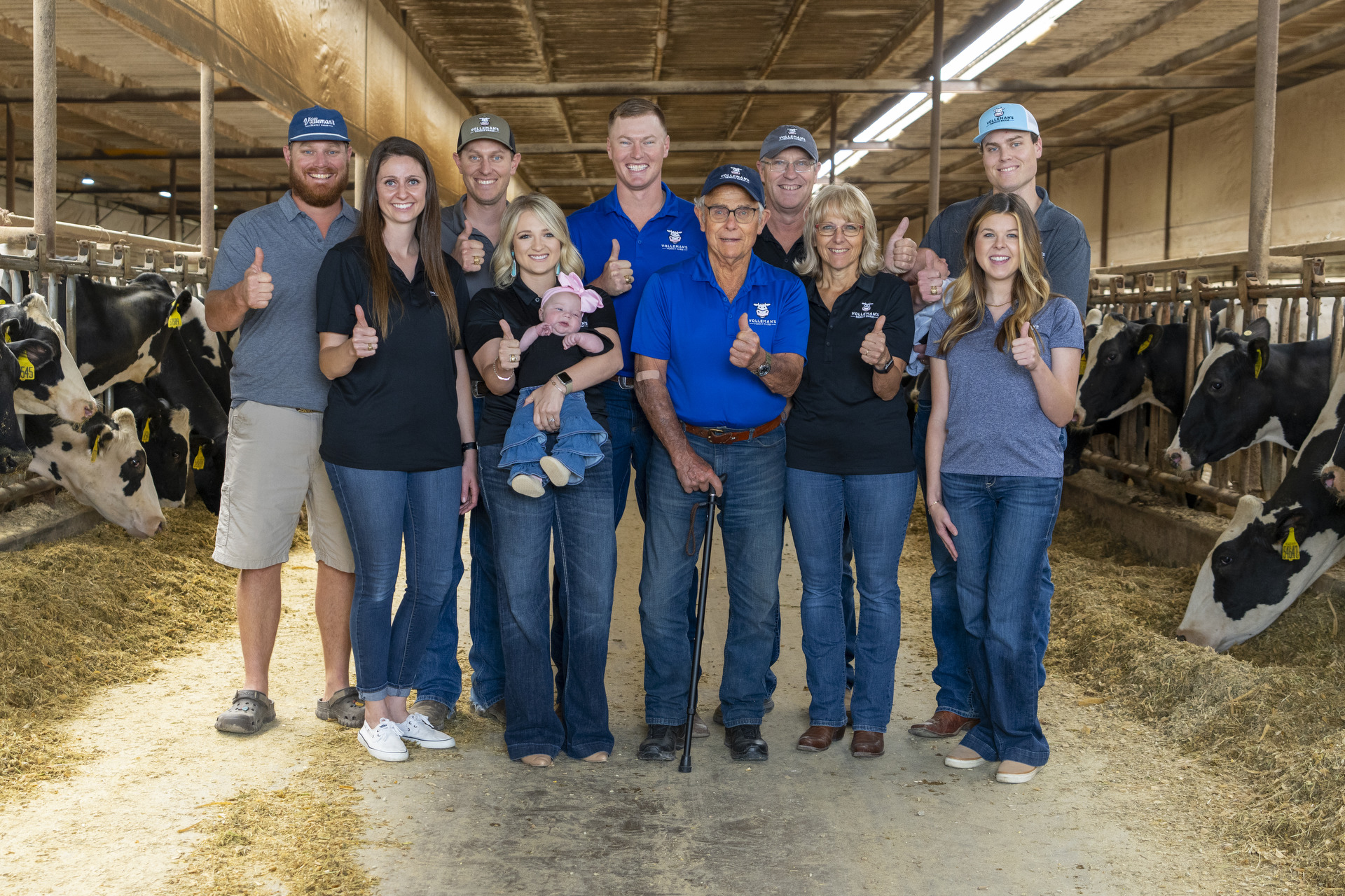 Members of the Volleman family, 6 men and 4 women, stand in the middle of the feed barn at the Volleman's Family Farm. They are all giving a thumbs up. 