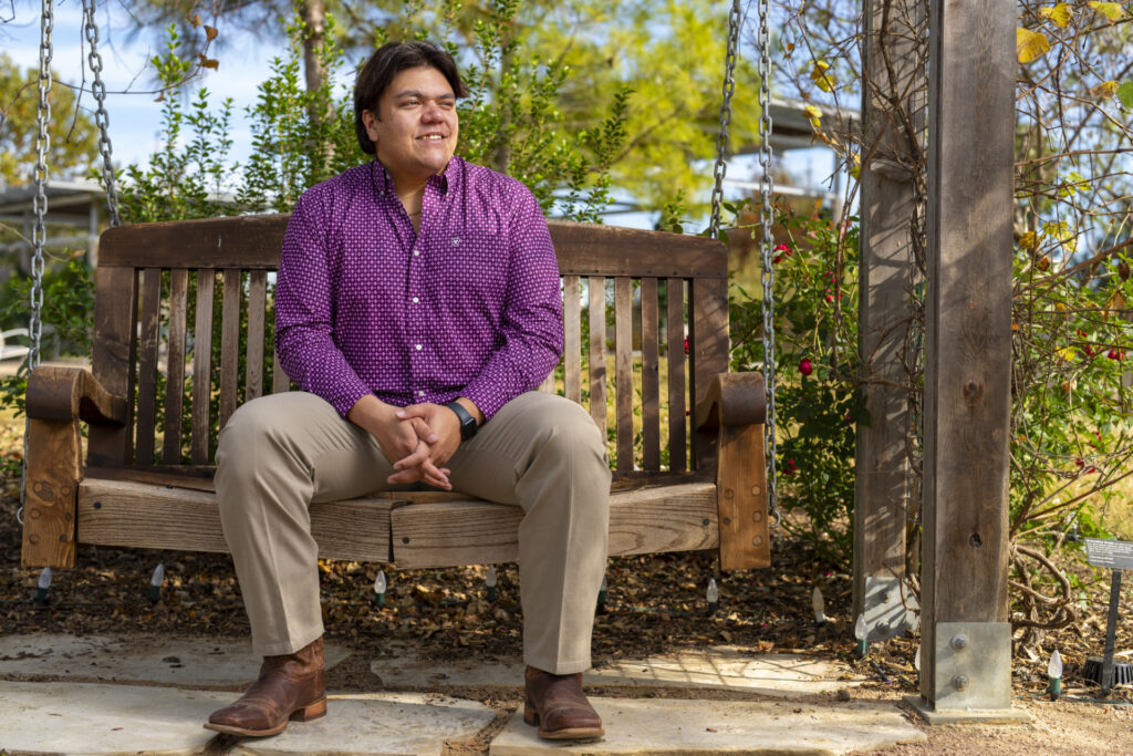 Dylan Sione sits on a bench swing looking off to his right. He is wearing a purple shirt, khakis and brown boots. There are plants and trees behind him. 