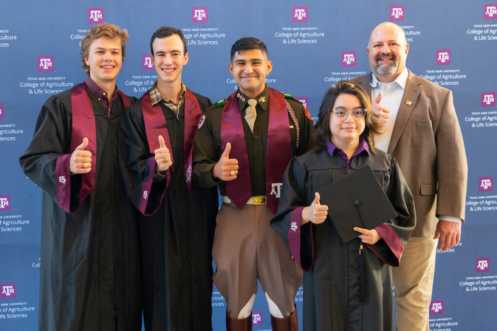 Four students wearing graduation robes give a thumbs up next to a man in a suit jacket also giving a thumbs up. The four are the first cohort to graduate from the Department of Rangeland, Wildlife and Fisheries Management.