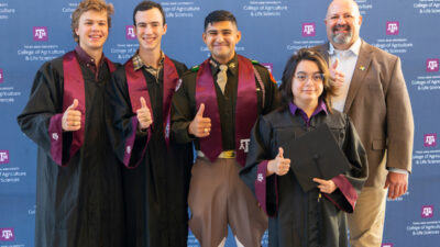 Four students in graduation robes give thumbs up next to a man in a suit jacket also giving thumbs up.