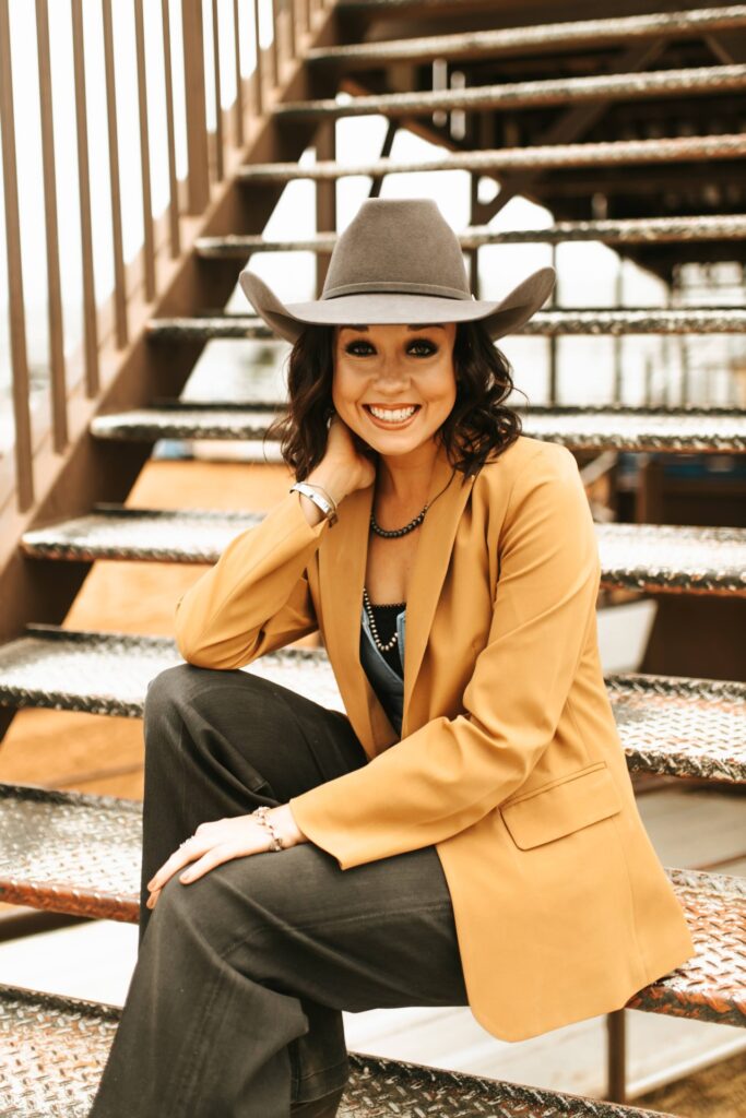 Hannah Crandall sits on a staircase. She weara camel colored balzer and beige cowboy hat.