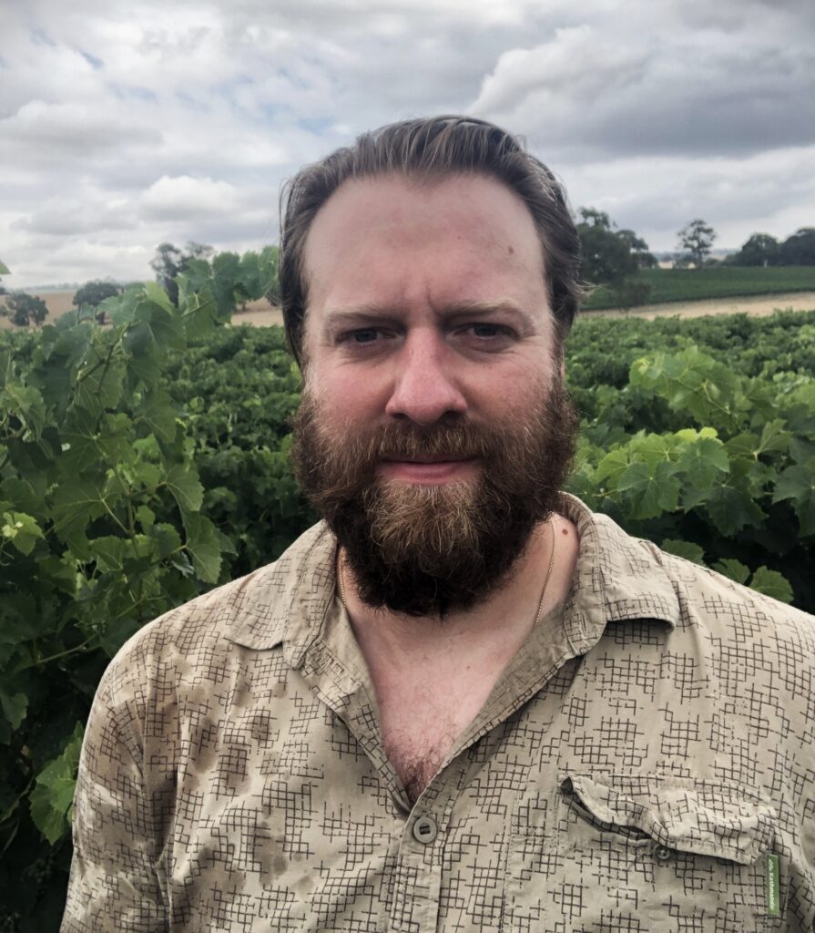 A head and shoulder picture of a man, Patrick O'Brien, Ph.D., who is a  viticulture specialist. He has green crops behind him and he is wearing a tan shirt.