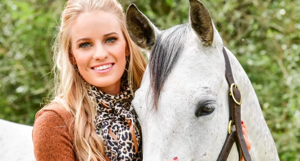 Kelly Ranly poses next to a white horse. The cowgirl has long blonde hair and wears an animal print neckerchief.