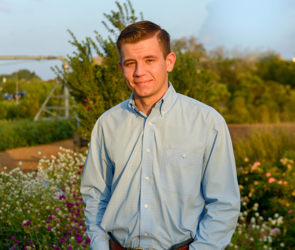 A smiling student, John Reaves, stands in a flower garden. He is wearing a long sleeve blue shirt. 