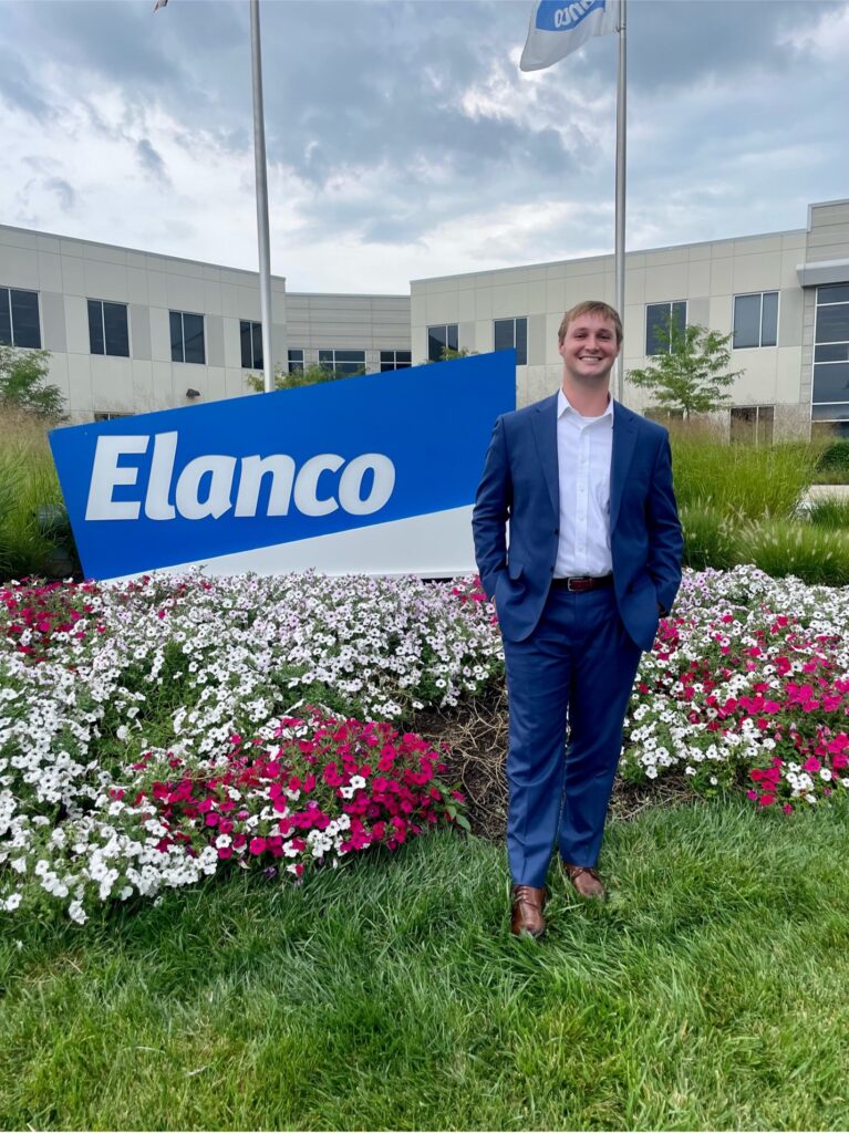 A full body image of poultry science student Ryder Nielson wearing a suit while standing in front of a flower bed with a sign behind him that reads "Elanco". 
