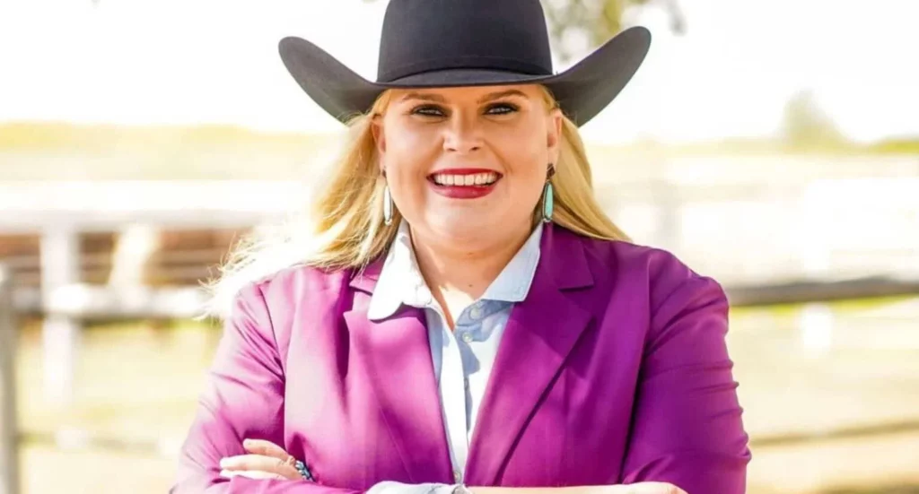 Cowgirl Tyler Schuster stands with arms crossed smiling atthe camera. She wears a black cowboy hot and fuschia blazer.
