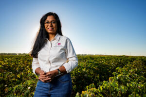A woman, Nithya Rajan, Ph.D., stands in a field. She is wearing blue jeans and a white long sleeve shirt. 