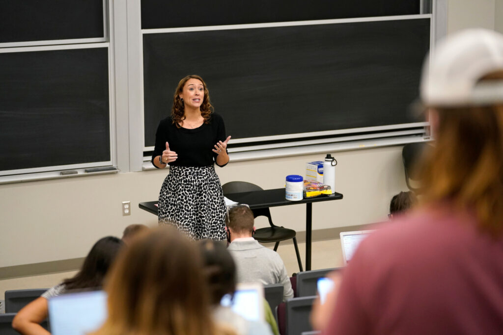 Jennifer, Strong, Ph.D. wear a black top ad black and white skirt. She lectures in a Texas A&M classromm to a large group of students.