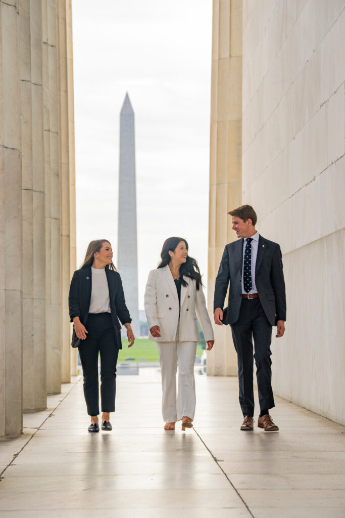 Two young women and a man, all dressed in suits and part of the agricultural policy internships program, walk along a marble outdoor corridor with Washington D.C. building behind them. 
