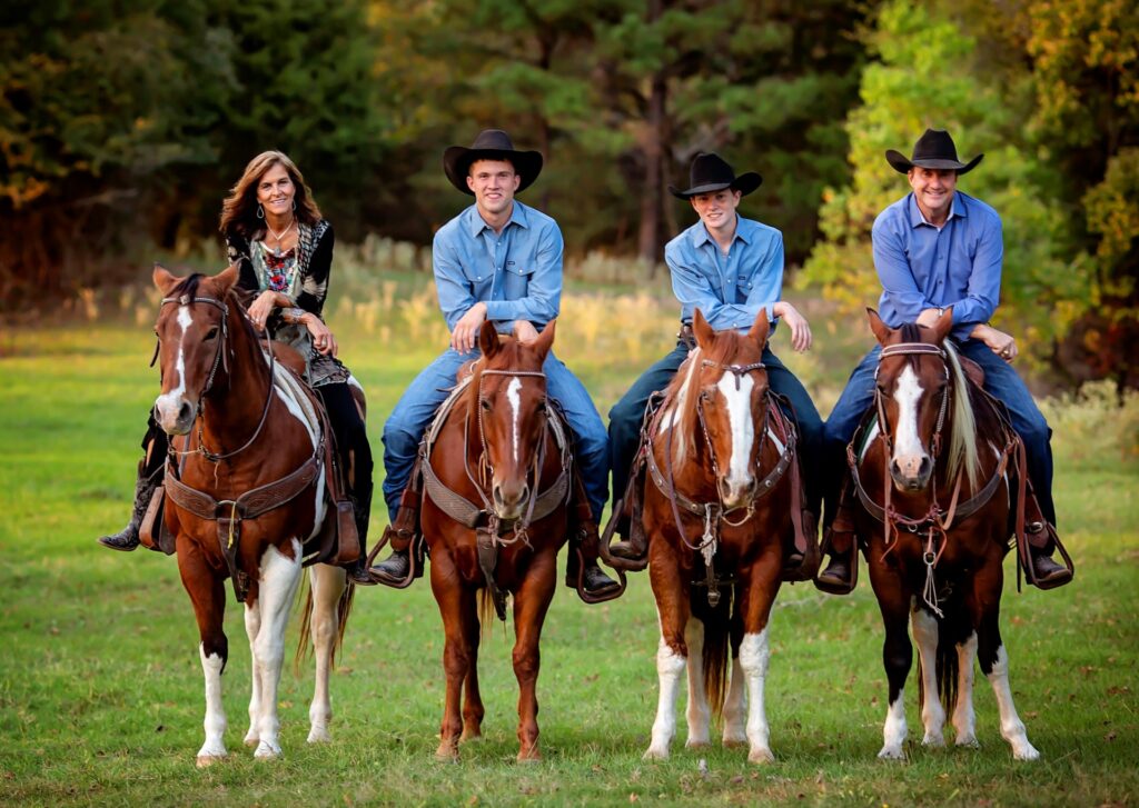 A woman and three men sitting atop horses. The woman is wearing a multi-colored dress. The men all have on blue shirts with blue jeans and a black cowboy hat.