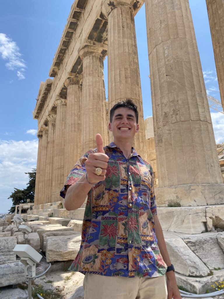 A young man, Daniel MacKenzie, in a floral shirt gives the thumbs up sign in front of the Acropolis in Greece.
