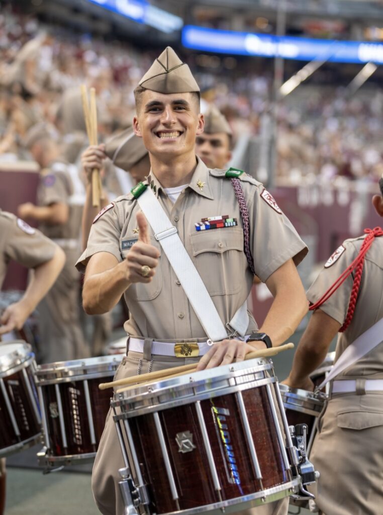 A young man, Daniel MacKenzie, in a Corps of Cadets uniform plays the drum at a Texas A&M football game.