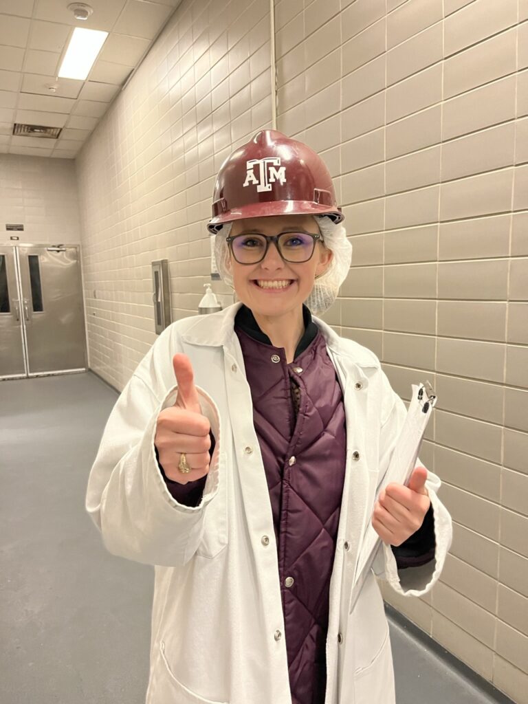 A woman, Katie Graves, stands in the hallway of a building with her meat judging lab coat on, a clip board in her hand, a hard hat with Texas A&M's logo and a thumbs up
