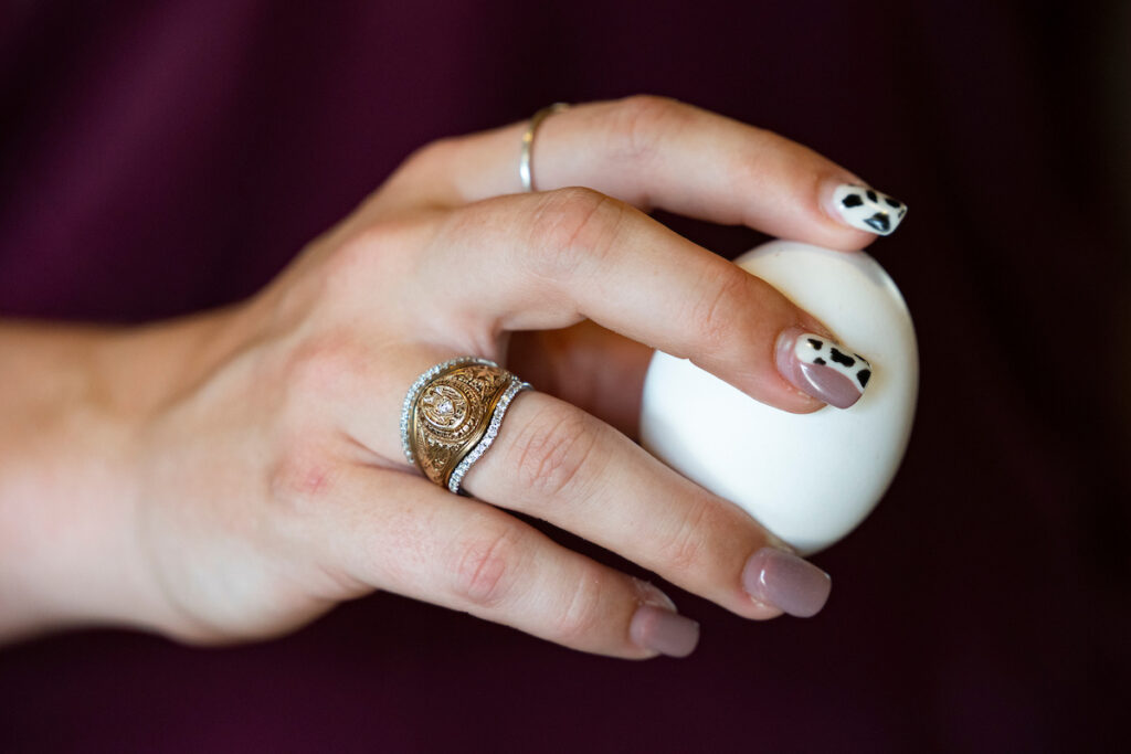 A woman's hand holding a white egg. Her nails are elaborately decorated and she wears a Texas A&M ring.
