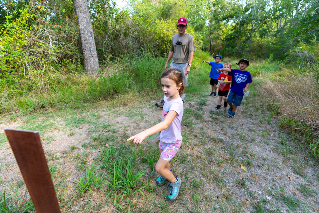 A girl points to a sign with three boys in the background pointing to something in the forest. 