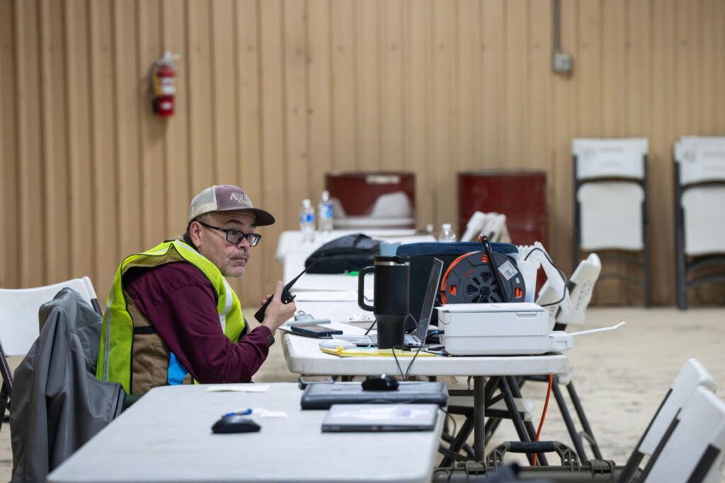 Richie Griffin sits in his yellow emergency vest with a walkie talkie in his hand in front of other equipment on tables as a part of the wildfire disaster response animal supply point