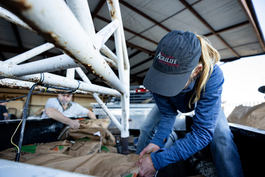 a woman in a Texas A&M AgriLife Extension cap loads feed at an animal supply point 