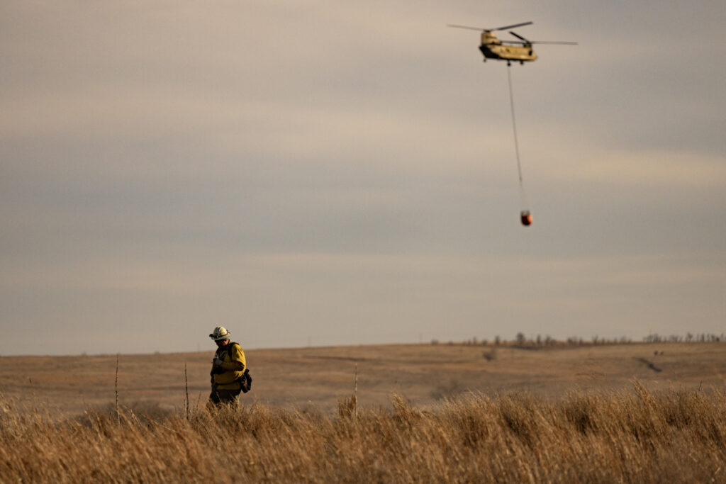 A Chinook helicopter flies a bucket of water to the flames as a firefighter walks in tall grass along a fence line.