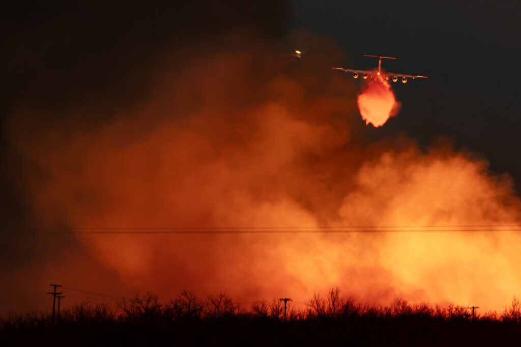 A wall of orange flames and smoke reach into the night sky as a firefighting plane releases a load of water that appears orange from the reflection of the flames
