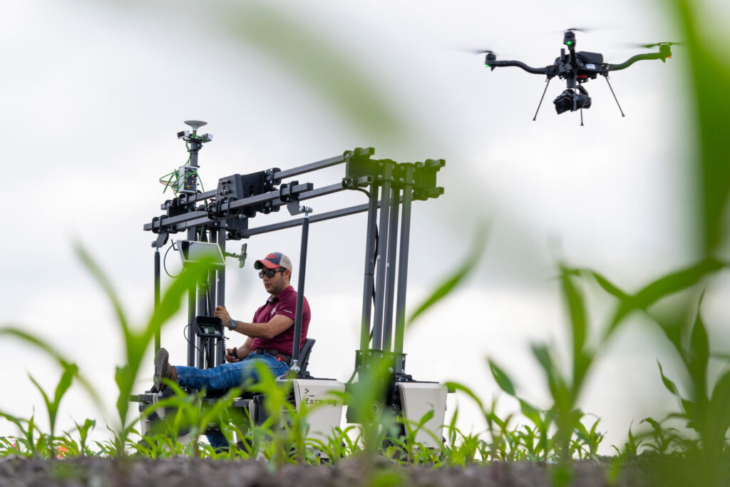 a man rides on a machine loaded with sensors, cameras and other technology - the tools of artificial intelligence - that is moving through a field of young corn plants with a drone hovering above it.