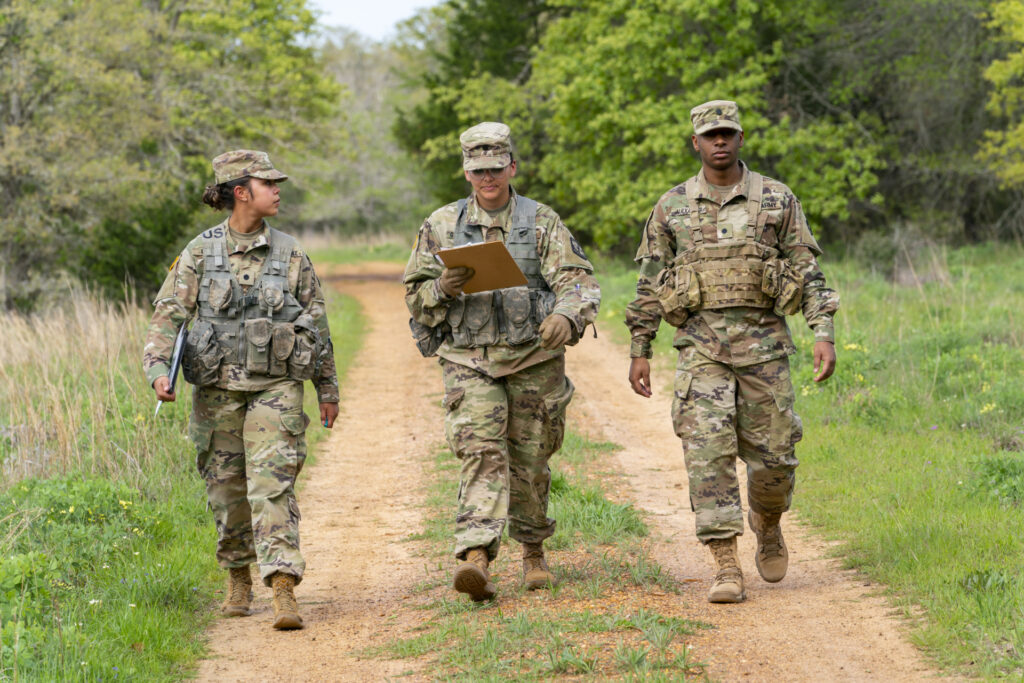 Three members of the Texas A&M Corps of Cadets walking on a dirt road with trees behind them. They are all wearing camouflage. 