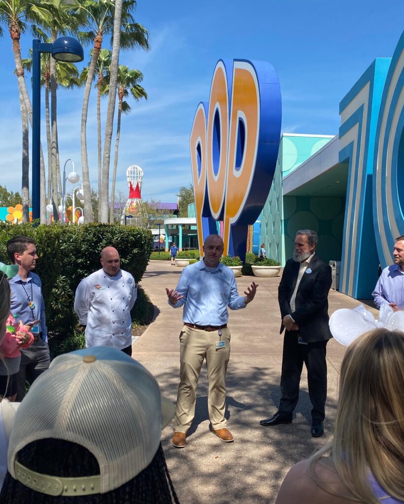 Five men, Disney "cast members" speaking to students. There is a large display that says POP behind them 