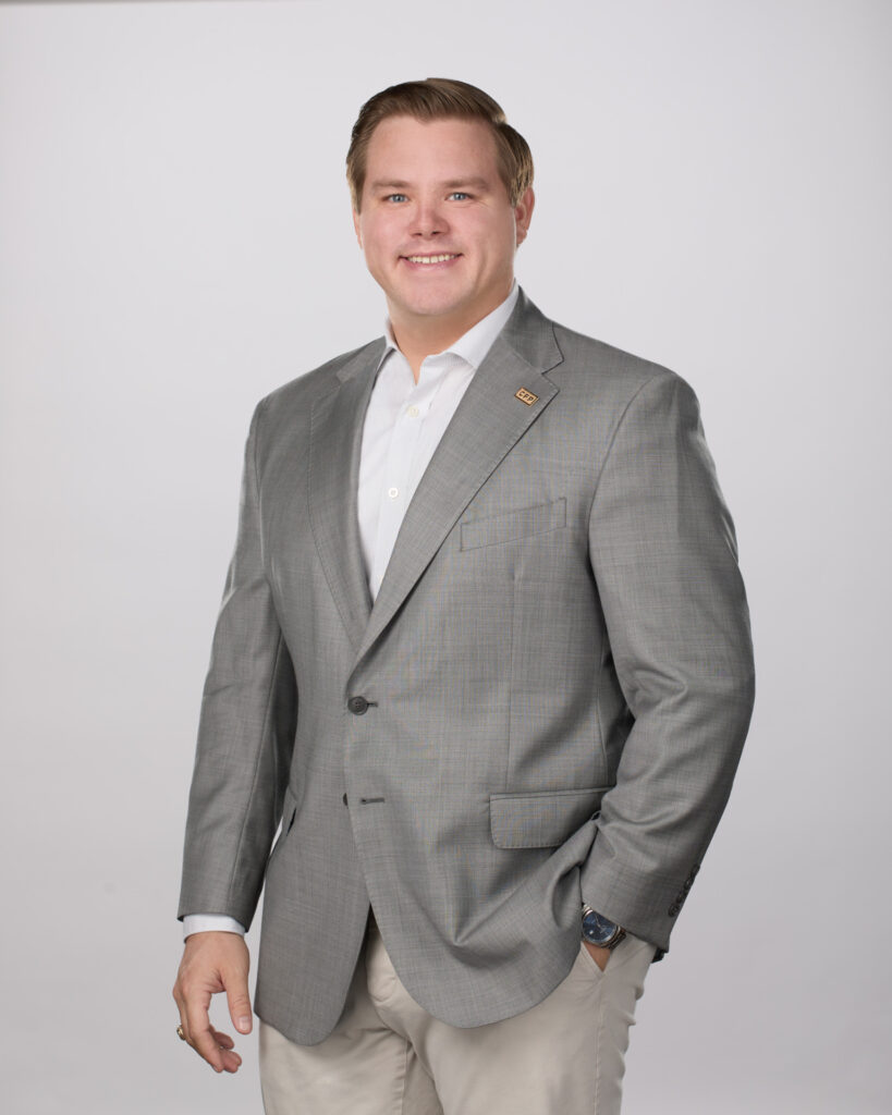 Photo of Riley Woodruff, certified financial planner. He is wearing a white shirt with a gray jacket and light colored pants. He has his left hand in his pocket. 