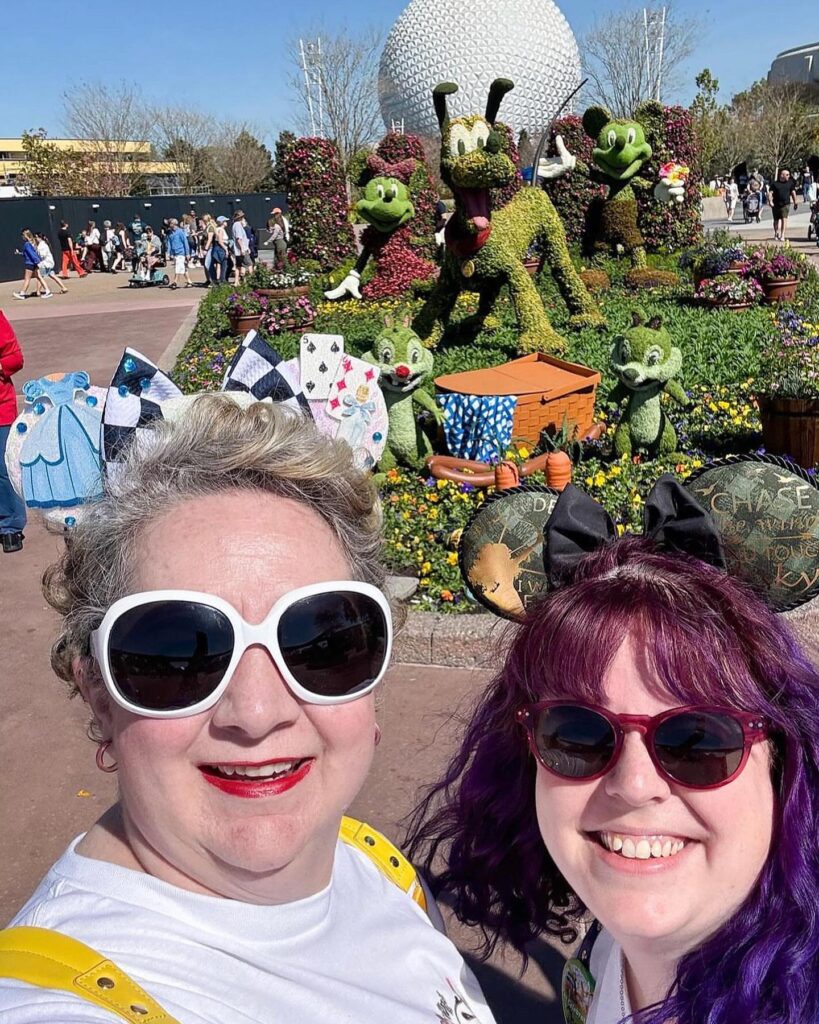 Two women, Dr. Heather Eden and Melyssa-Anne Stricklin, taking a selfie at Disney World, The Spaceship Earth globe is behind them as well as four bushes shaped as Chip 'n' Dale,  Pluto and Minnie Mouse. 