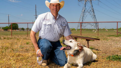 A man wearing a cowboy hat kneels next to a white livestock guardian dog who wears a GPS collar.