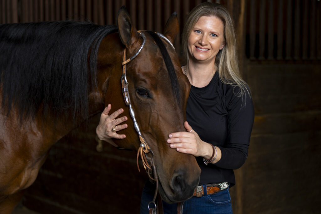 A woman, Meredith Neely, stand with a horse. She is wearing a black top with blue jeans. The horse is brown with a dark mane. 