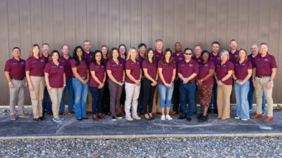 Group of 24 people wearing matching maroon shirts standing outside a building for a group picture.