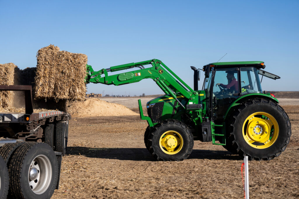a green tractor with hay forks on it unloads square hay bales at an Animal Supply Point