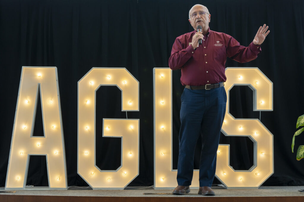 A man in a maroon button down and navy pants speaks with a microphone on a stage in front of the letters "AGLS" that are illuminated by small light bulbs.