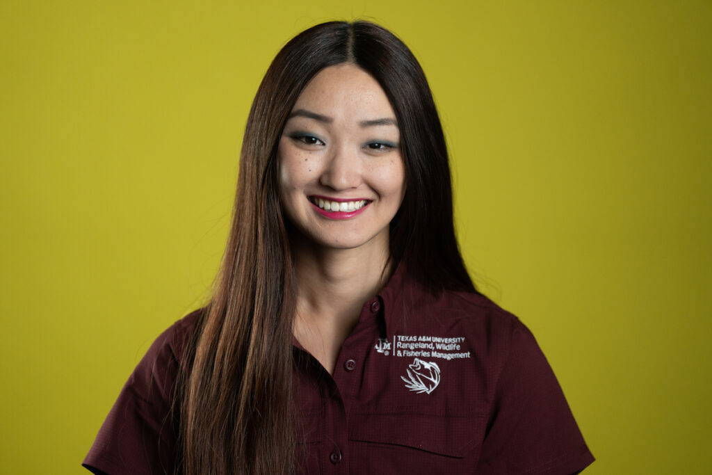 A headshot of Hannah Evans, a young woman with long dark hair, wearing a maroon Aggie shirt agaisnt a chartreuse background