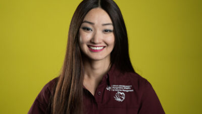 A headshot of a young woman with long dark hair wearing a maroon shirt agaisnt a chartreuse background