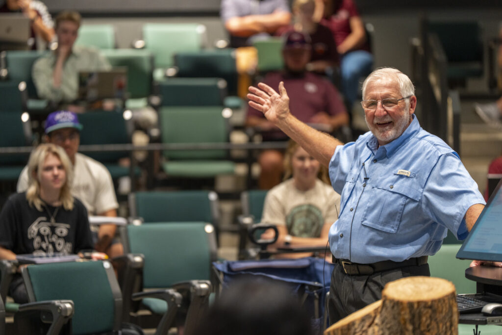 David Reed, Ph.D. wears a light blue, short sleeve button-up shirt while lecturing for Horticultural Science and Practices with his right hand in the air.