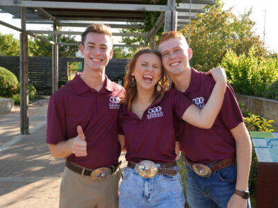 Three people in maroon polos stand close together with one person holding their thumb up.