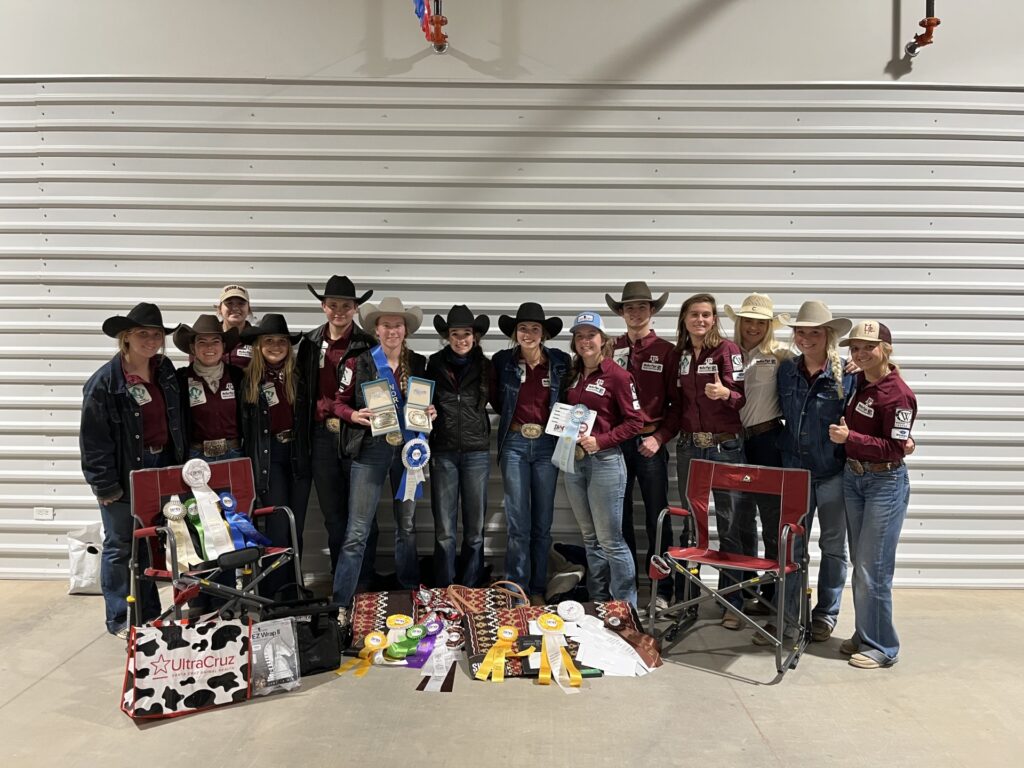 A team of students dressed in maroon shirts and western cowboy hats have a collection of ribbons, chairs, bags and plaques laid out in front of them after winning 