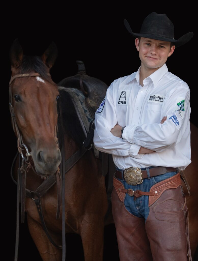 A young man, student Tucker Gillespie, stands beside his horse with his arms folded. He is dressed in a white shirt with logo patches on it and a black cowboy hat