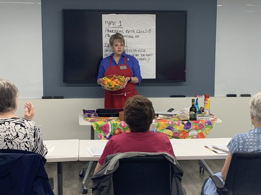 A Master Wellness Volunteer in a blue shirt, wearing a red apron and holding a bowl of vegetables. speaks to three women who are seated in front of her.