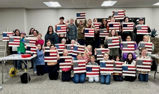 A large group photo of individuals holding up squares for a quilt. The squares are replicas of the U.S. flag and are red white and blue. 