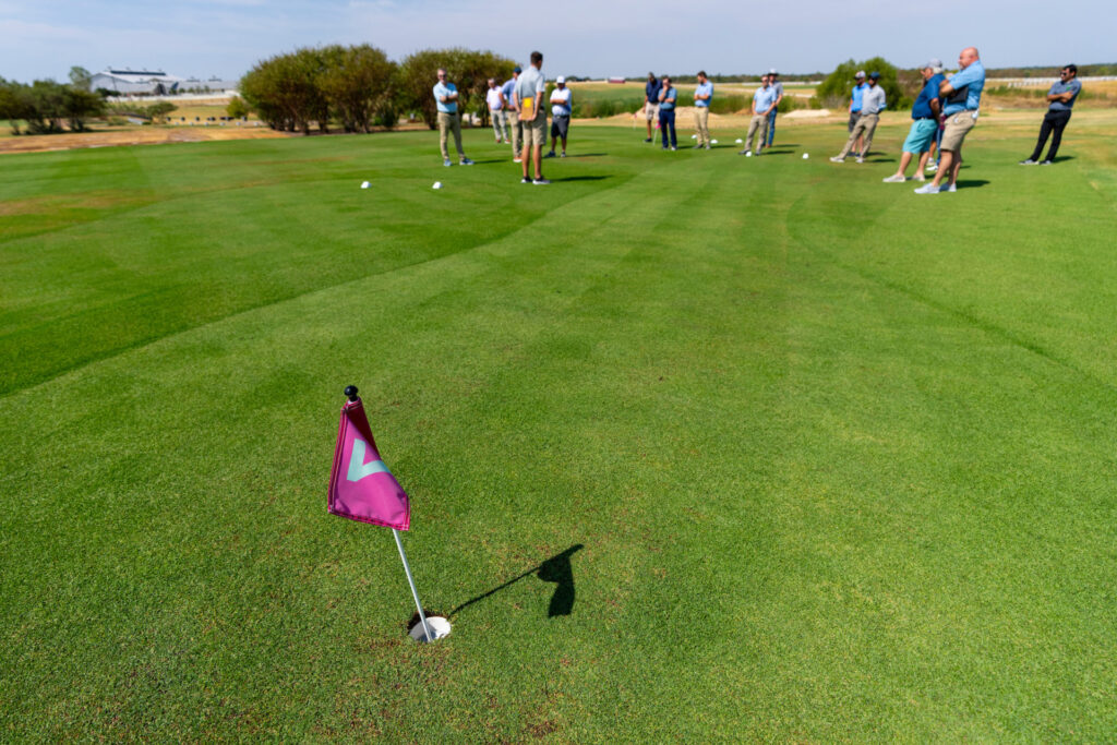 Several individuals standing on a putting green. There is a small maroon flag in a hole.