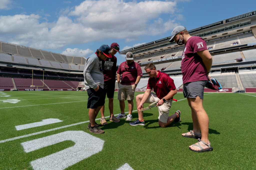 Men gathered around one man who is testing turfgrass sample inside Kyle Field at Texas A&M. 