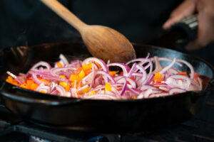 A person is cooking a dish with onions and bell peppers.