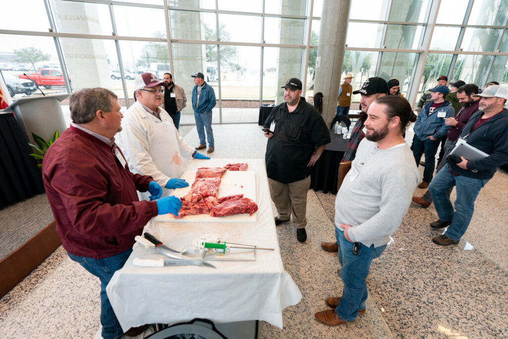 Two men, Ray Riley and Davey Griffin, Ph.D., stand at a table with uncooked briskets with a group of men standing around in a large glassed meeting room. 