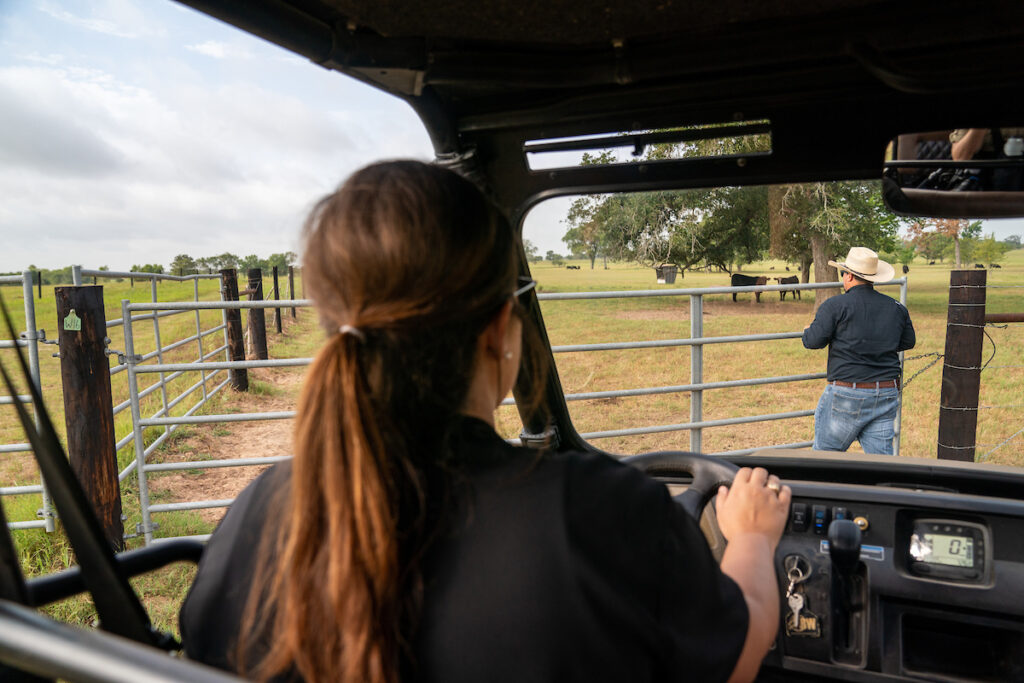 A woman sits at the wheel of an all-terrain vehicle as a man in a cowboy hat opens a gate to a field of cattle.  