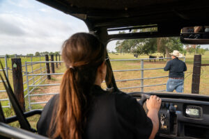 A woman sits at the wheel of an all-terrain vehicle as a man in a cowboy hat opens a gate to a field of cattle.
